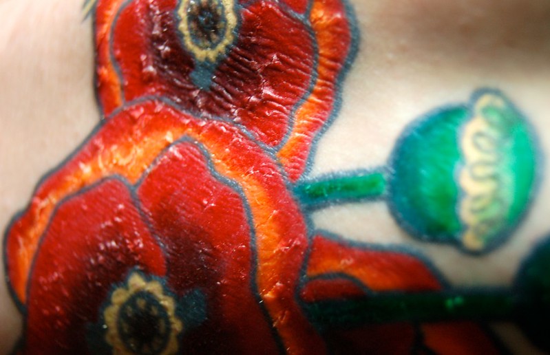 A close-up of a colorful tattoo depicting red poppy flowers with green stems and leaves on human skin. The tattoo is vibrant and shows detailed outlines and shading.